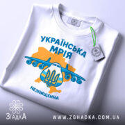 Футболка українська мрія, біла, складена на поверхні, фон світлий, принт з літаком та гербом України, кольори жовтий та блакитний, текст 'УКРАЇНСЬКА МРІЯ' та 'НЕЗНИЩЕННА', на ярлику зелена стрічка.