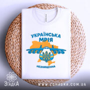 Футболка українська мрія, біла, складена на плетеній основі, фон нейтральний, принт з літаком та гербом України, кольори жовтий та блакитний, текст 'УКРАЇНСЬКА МРІЯ' та 'НЕЗНИЩЕННА'.