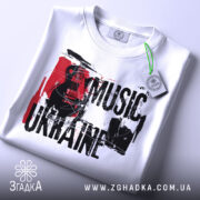 Футболка українська музика, біла, складена на столі, фон світло-сірий, принт з чорним текстом 'MUSIC UKRAINE' та гітарою, прикріплена етикетка з зеленим шнурком.