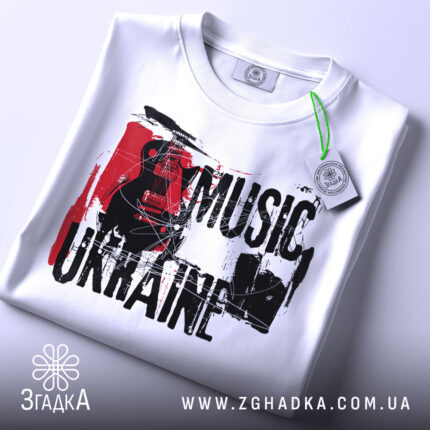 Футболка українська музика, біла, складена на столі, фон світло-сірий, принт з чорним текстом 'MUSIC UKRAINE' та гітарою, прикріплена етикетка з зеленим шнурком.
