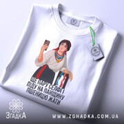 Футболка українська селфі красуня, біла, зображена з верхнього ракурсу, на фоні світло-сірого кольору, принт з дівчиною в національному вбранні, яка тримає телефон, на футболці є етикетка з логотипом бренду, текст під малюнком: 'ЩЕ ПАРУ СЕЛФІ ПІДУ НА ПАНЦІЮ ПШЕНИЦЮ ЖАТИ'.