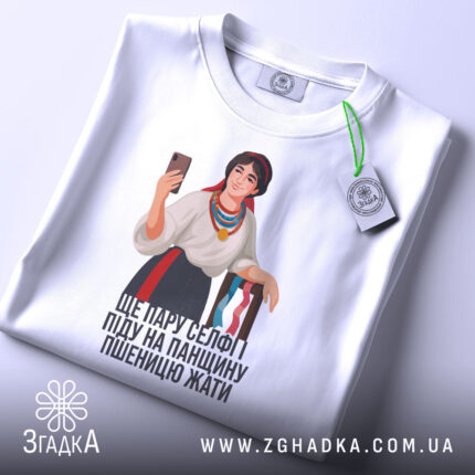 Футболка українська селфі красуня, біла, зображена з верхнього ракурсу, на фоні світло-сірого кольору, принт з дівчиною в національному вбранні, яка тримає телефон, на футболці є етикетка з логотипом бренду, текст під малюнком: 'ЩЕ ПАРУ СЕЛФІ ПІДУ НА ПАНЦІЮ ПШЕНИЦЮ ЖАТИ'.