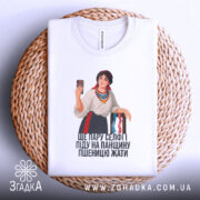 Футболка українська селфі красуня, біла, зображена з верхнього ракурсу, на фоні плетеного кошика, принт з дівчиною в національному вбранні, яка тримає телефон, текст під малюнком: 'ЩЕ ПАРУ СЕЛФІ ПІДУ НА ПАНЦІЮ ПШЕНИЦЮ ЖАТИ'.
