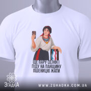 Футболка українська селфі красуня, біла, зображена з верхнього ракурсу, на фоні світло-сірого кольору, принт з дівчиною в національному вбранні, яка тримає телефон, на футболці є етикетка з логотипом бренду, текст під малюнком: 'ЩЕ ПАРУ СЕЛФІ ПІДУ НА ПАНЦІЮ ПШЕНИЦЮ ЖАТИ'.