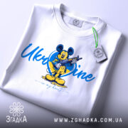 Футболка український дух, біла, зображена на столі, фон світлий, принт з мультяшним персонажем, на якому видно етикетку, напис 'Ukraine in my heart'.