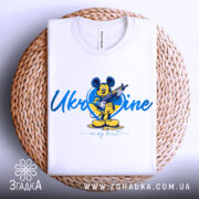 Футболка український дух, біла, зображена на плетеній основі, фон світлий, принт з мультяшним персонажем, напис 'Ukraine in my heart'.