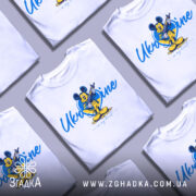 Футболка український дух, біла, зображена на манекені, фон градієнтний сірий, принт з мультяшним персонажем у жовтій та блакитній кольоровій гамі, напис 'Ukraine in my heart'.