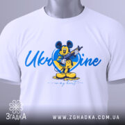 Футболка український дух, біла, зображена на столі, фон світлий, принт з мультяшним персонажем, на якому видно етикетку, напис 'Ukraine in my heart'.