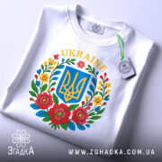 Футболка український герб, біла, зображена зверху, складена на поверхні, фон світло-сірий, принт містить герб України в синьому квадраті оточений кольоровими квітами, на комірці видна етикетка.
