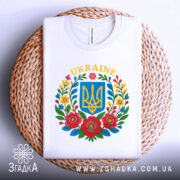 Футболка український герб, біла, зображена зверху, складена на плетеній основі, фон світло-сірий, принт містить герб України в синьому квадраті оточений кольоровими квітами.