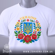 Футболка український герб, біла, зображена зверху, складена на поверхні, фон світло-сірий, принт містить герб України в синьому квадраті оточений кольоровими квітами, на комірці видна етикетка.