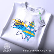 Футболка український хіпстер, біла, зображена зверху, фон світлий, принт з персонажем у жовто-блакитному вбранні, напис "100% українським хіпстер", на футболці є етикетка з зеленим шнурком.