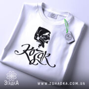 Футболка український козак, білого кольору, складена на поверхні, фон світло-сірий, принт з чорним малюнком козака та написом 'Kozak', на футболці є етикетка з зеленим шнурком.