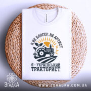 Футболка український тракторист, білого кольору, зображена на плетеній основі, фон світло-сірий, принт з трактором, сонцем і рослиною, текст чорного кольору.