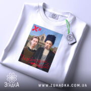 Футболка український вайб, біла, складена на столі, фон світлий, принт зображає двох людей у традиційному одязі, текст "У український вайб" червоним кольором, на футболці є етикетка з логотипом.