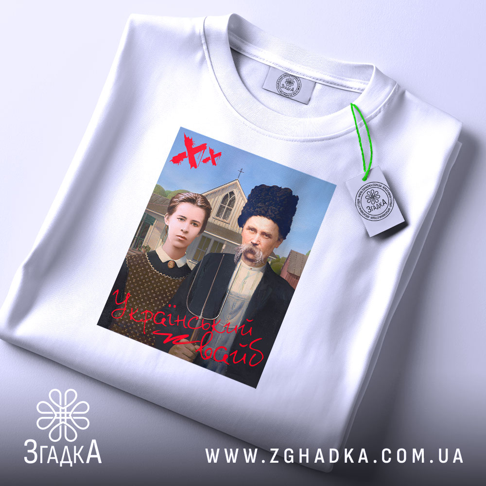 Футболка український вайб, біла, складена на столі, фон світлий, принт зображає двох людей у традиційному одязі, текст "У український вайб" червоним кольором, на футболці є етикетка з логотипом.