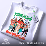 Футболка улюблений вихователь, білого кольору, зображена на столі, ракурс зверху, фон світлий, принт з персонажами, що святкують, з яскравими кольорами та етикеткою на комірці.