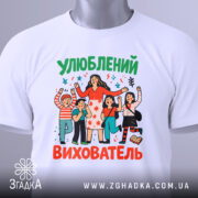 Футболка улюблений вихователь, білого кольору, зображена на столі, ракурс зверху, фон світлий, принт з персонажами, що святкують, з яскравими кольорами та етикеткою на комірці.