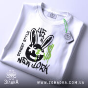 Футболка урбан стиль, біла, зображена зверху, фон світлий, принт з чорним смайлом з вухами зайця, зеленими краплями фарби та написом 'NEW YORK', на комірці видна етикетка.