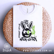 Футболка урбан стиль, біла, зображена зверху на плетеній основі, фон світлий, принт з чорним смайлом з вухами зайця, зеленими краплями фарби та написом 'NEW YORK'.