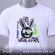 Футболка урбан стиль, біла, зображена зверху, фон світлий, принт з чорним смайлом з вухами зайця, зеленими краплями фарби та написом 'NEW YORK', на комірці видна етикетка.