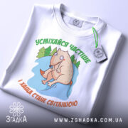 Футболка усміхайся частіше, білого кольору, складена на столі, фон світло-сірий, принт з веселим персонажем, що сидить на камені серед дерев, текст зеленого кольору зверху та оранжевого знизу, з етикеткою на рукаві.