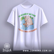 Футболка усміхайся частіше, білого кольору, підвішена на вішалці, фон світло-сірий, принт з веселим персонажем, що сидить на камені серед дерев, текст зеленого кольору зверху та оранжевого знизу.