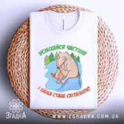 Футболка усміхайся частіше, білого кольору, складена на плетеній кошику, фон світло-сірий, принт з веселим персонажем, що сидить на камені серед дерев, текст зеленого кольору зверху та оранжевого знизу.