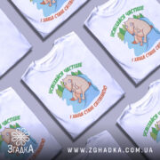 Футболка усміхайся частіше, білого кольору, зображена на манекені, фон світло-сірий, принт з веселим персонажем, що сидить на камені серед дерев, текст зеленого кольору зверху та оранжевого знизу.