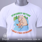 Футболка усміхайся частіше, білого кольору, складена на столі, фон світло-сірий, принт з веселим персонажем, що сидить на камені серед дерев, текст зеленого кольору зверху та оранжевого знизу, з етикеткою на рукаві.