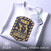 Футболка увага про роботу, біла, складена на поверхні, фон світло-сірий, принт у жовто-синіх тонах з написом 'УВАГА' та ілюстрацією двох фігур, з етикеткою на рукаві.