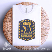 Футболка увага про роботу, біла, складена на плетеній основі, фон світло-сірий, принт у жовто-синіх тонах з написом 'УВАГА' та ілюстрацією двох фігур.