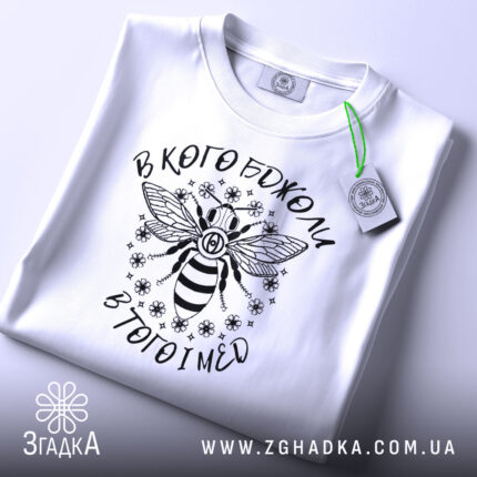 Футболка в кого бджоли, біла, зображена зверху, складена з етикеткою, фон світлий, принт з бджолою та написом вгорі і внизу.