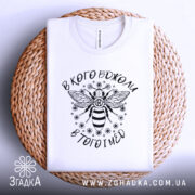 Футболка в кого бджоли, біла, складена на плетеній кошику, фон світлий, принт з бджолою та написом вгорі і внизу.