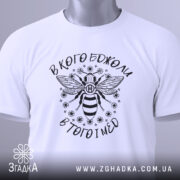 Футболка в кого бджоли, біла, зображена зверху, складена з етикеткою, фон світлий, принт з бджолою та написом вгорі і внизу.