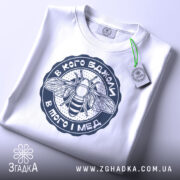 Футболка в кого бджоли, біла, складена на поверхні, з видимим етикеткою, фон світлий, принт з бджолою та написом у темно-синьому кольорі.