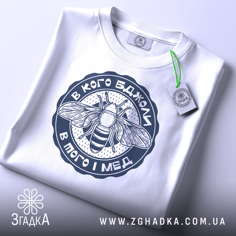 Футболка в кого бджоли, біла, складена на поверхні, з видимим етикеткою, фон світлий, принт з бджолою та написом у темно-синьому кольорі.