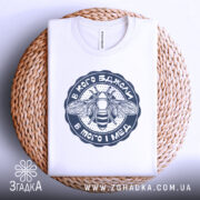 Футболка в кого бджоли, біла, складена на плетеній кошику, фон світлий, принт з бджолою та написом у темно-синьому кольорі.