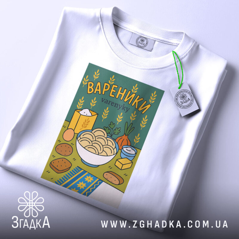 Футболка вареники білого кольору, складена на поверхні, з принтом вареників у мисці на зеленому фоні, прикрашена елементами, такими як цукор і овочі, з етикеткою, що звисає з коміра.