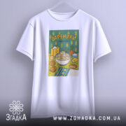 Футболка вареники білого кольору, підвішена на вішалці, з принтом вареників у мисці на зеленому фоні, деталі принта включають різні інгредієнти, розташовані навколо.