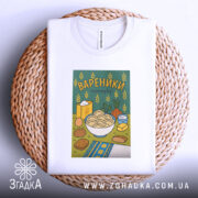 Футболка вареники білого кольору, складена на плетеній кошику, з принтом вареників у мисці на зеленому фоні, оточена елементами, такими як цукор і овочі, що додають кольору.