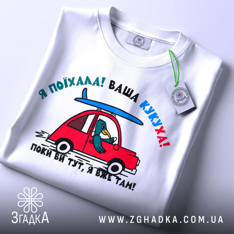 Футболка ваша кукуха, біла футболка, зображена зверху, з принтом червоного автомобіля та птаха, що сидить за кермом, фон світло-сірий, на футболці є етикетка.