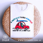 Футболка ваша кукуха, біла футболка, складена на плетеному кошику, з принтом червоного автомобіля та птаха, що сидить за кермом, фон світлий, принт яскравий і кольоровий.