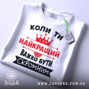 Футболка важко бути скромним. Біла футболка, складена на поверхні, зображена зверху. На фоні світло-сірий градієнт. Принт чорного та червоного кольорів з текстом та короною, з етикеткою на рукаві.