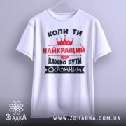 Футболка важко бути скромним. Біла футболка з коротким рукавом, зображена з переднього ракурсу на вішалці. На фоні світло-сірий градієнт. Принт чорного та червоного кольорів з текстом та короною.