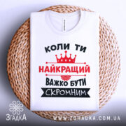 Футболка важко бути скромним. Біла футболка, складена на плетеній основі, зображена зверху. На фоні світло-сірий градієнт. Принт чорного та червоного кольорів з текстом та короною.