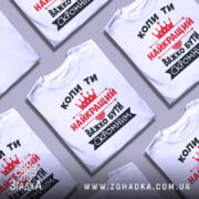 Футболка важко бути скромним. Біла футболка з коротким рукавом, зображена з правого ракурсу на манекені. На фоні світло-сірий градієнт. Принт чорного та червоного кольорів з текстом та короною.