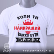 Футболка важко бути скромним. Біла футболка, складена на поверхні, зображена зверху. На фоні світло-сірий градієнт. Принт чорного та червоного кольорів з текстом та короною, з етикеткою на рукаві.