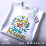 Футболка вчу серцем, білого кольору, зображена зверху, на плоскій поверхні, фон світло-сірий, принт з написом 'ВЧУ СЕРЦЕМ І РОЗУМОМ' в яскравих кольорах, з етикеткою на комірці.