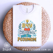 Футболка вчу серцем, білого кольору, зображена зверху на плоскій поверхні з плетеного матеріалу, фон світло-сірий, принт з написом 'ВЧУ СЕРЦЕМ І РОЗУМОМ' в яскравих кольорах.